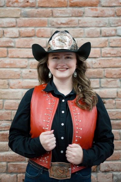 Photo of 2026 Queen Aubrey in black hat with medallion, black oxford, and red leather vest